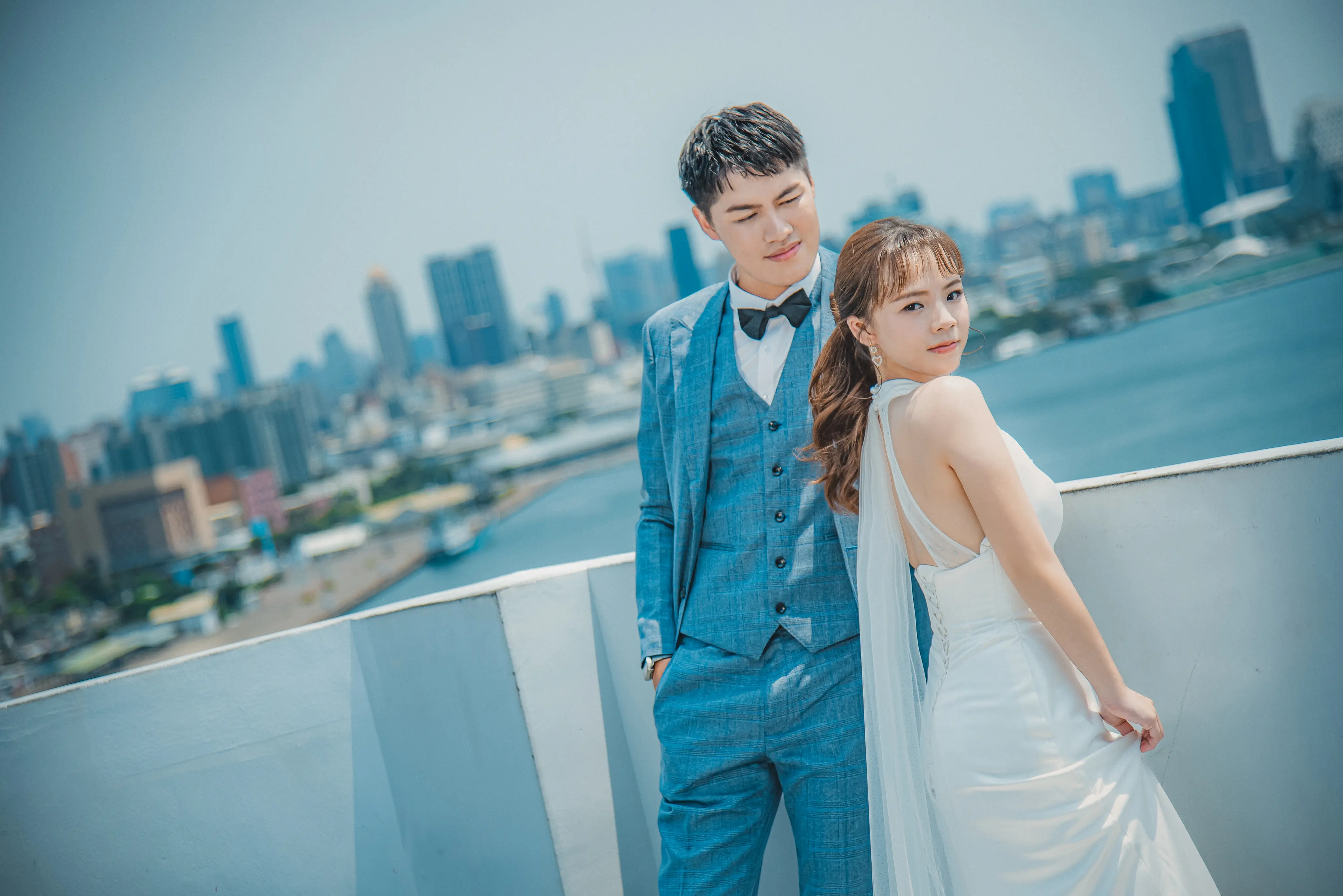 城市婚紗寫真：新人穿藍色西裝與白紗，站在海岸欄杆前合影