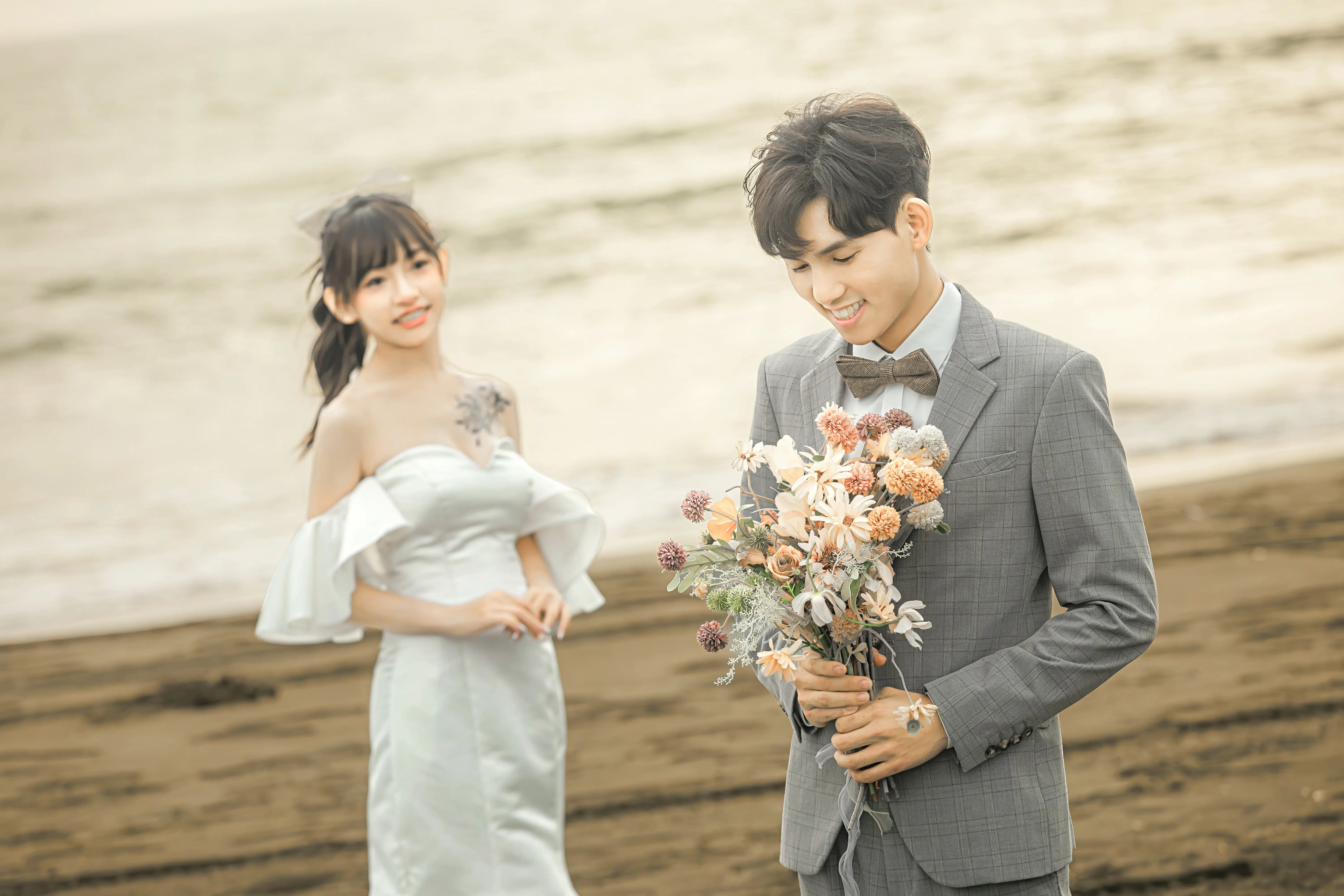 男生手持捧花低頭微笑，女生站在後方的海邊