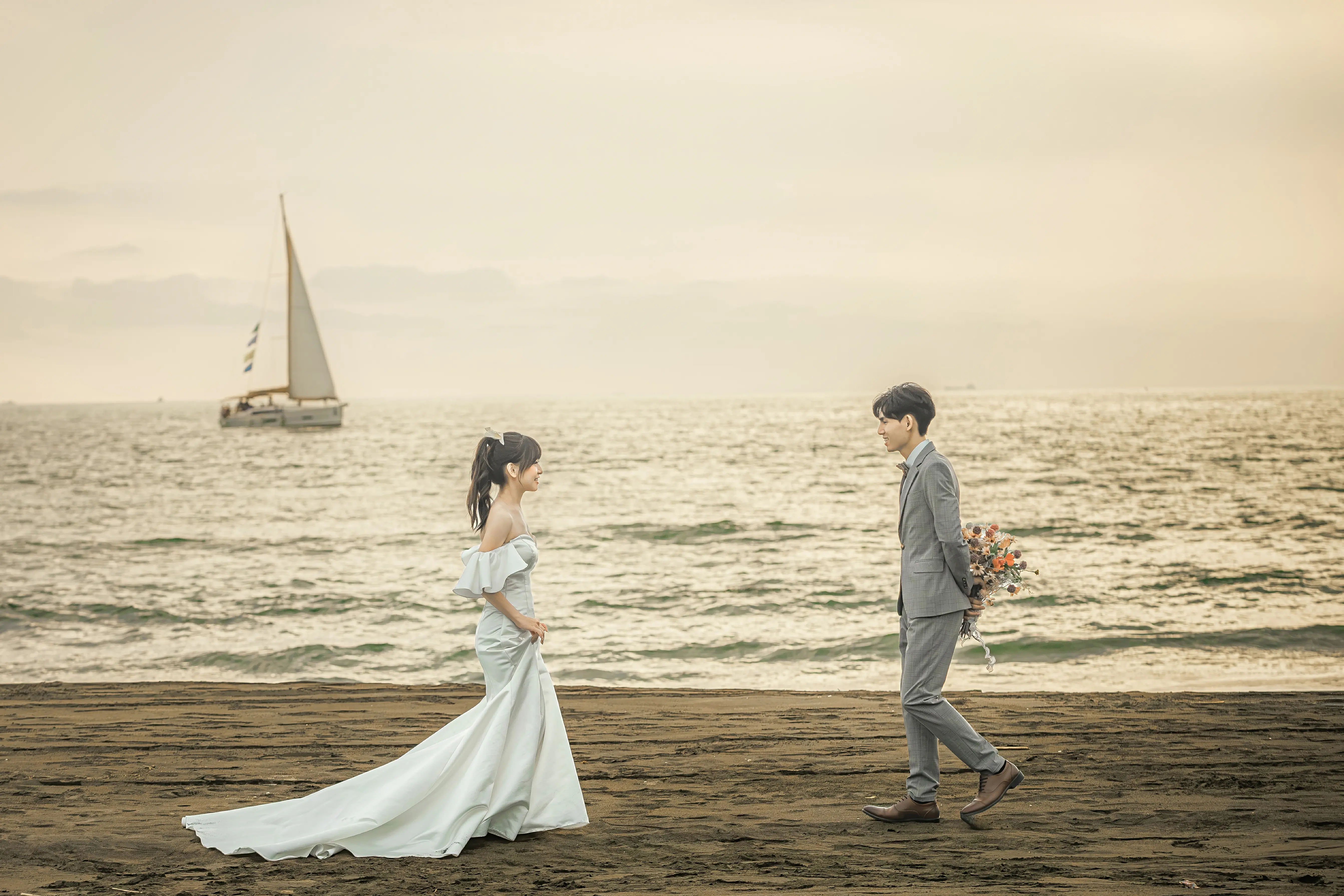 女生穿魚尾禮服與男生在海邊相向而行，後方海面有帆船