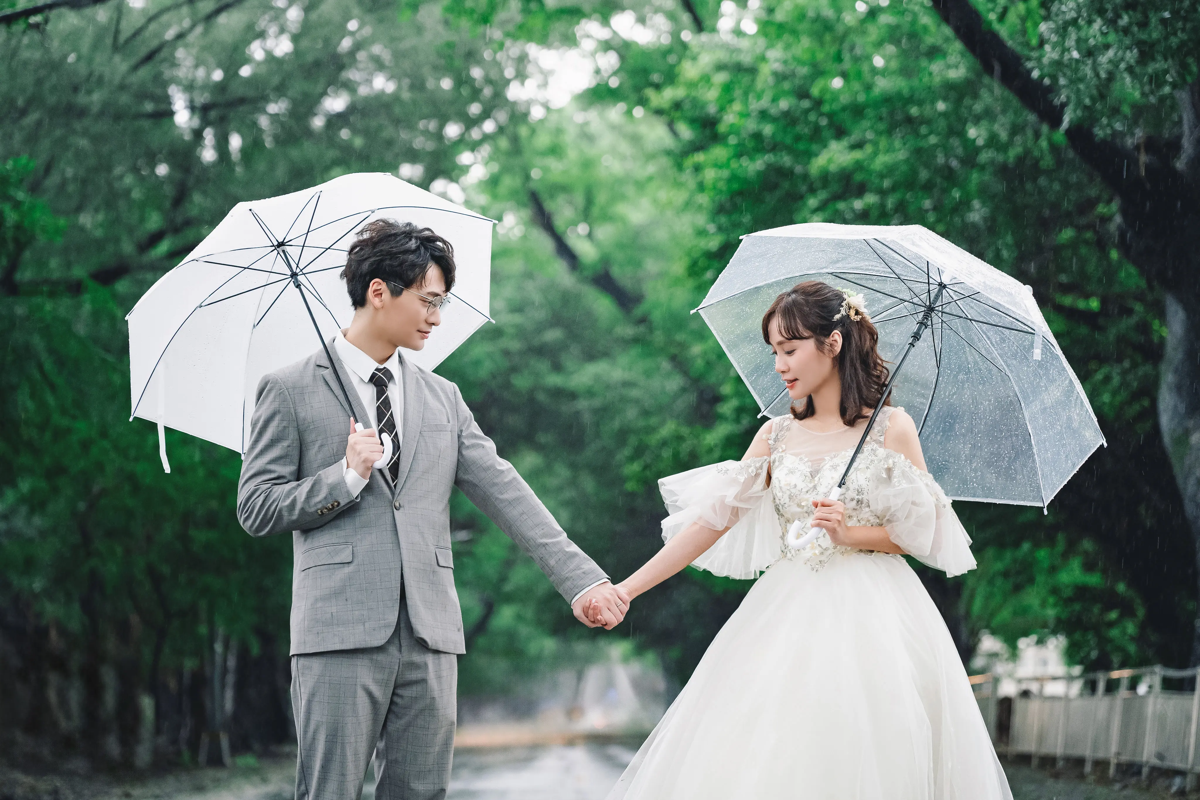 新人在綠樹步道上各撐透明雨傘，牽著手看向彼此
