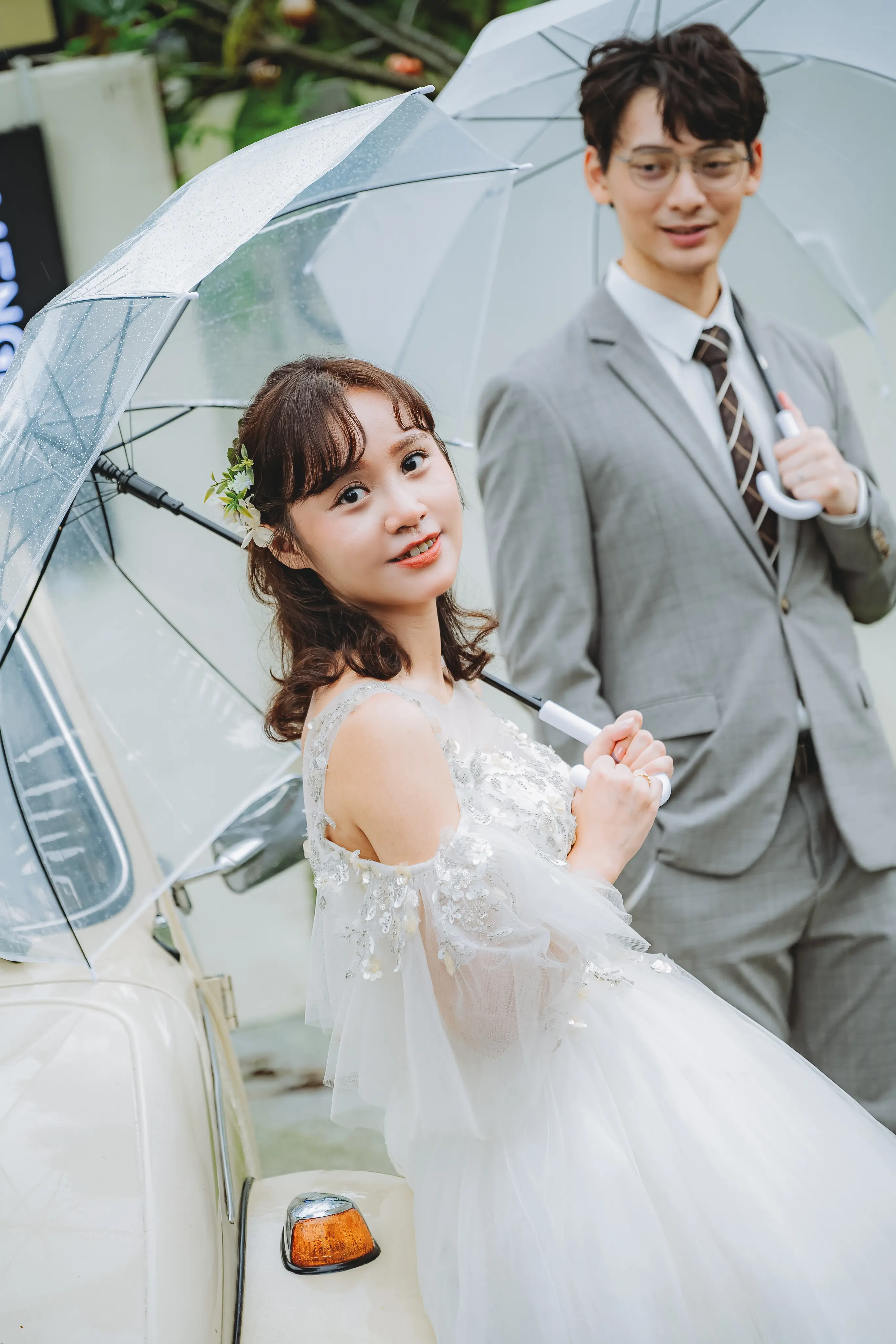 新娘在復古白色汽車旁撐傘回望鏡頭，新郎站在身後